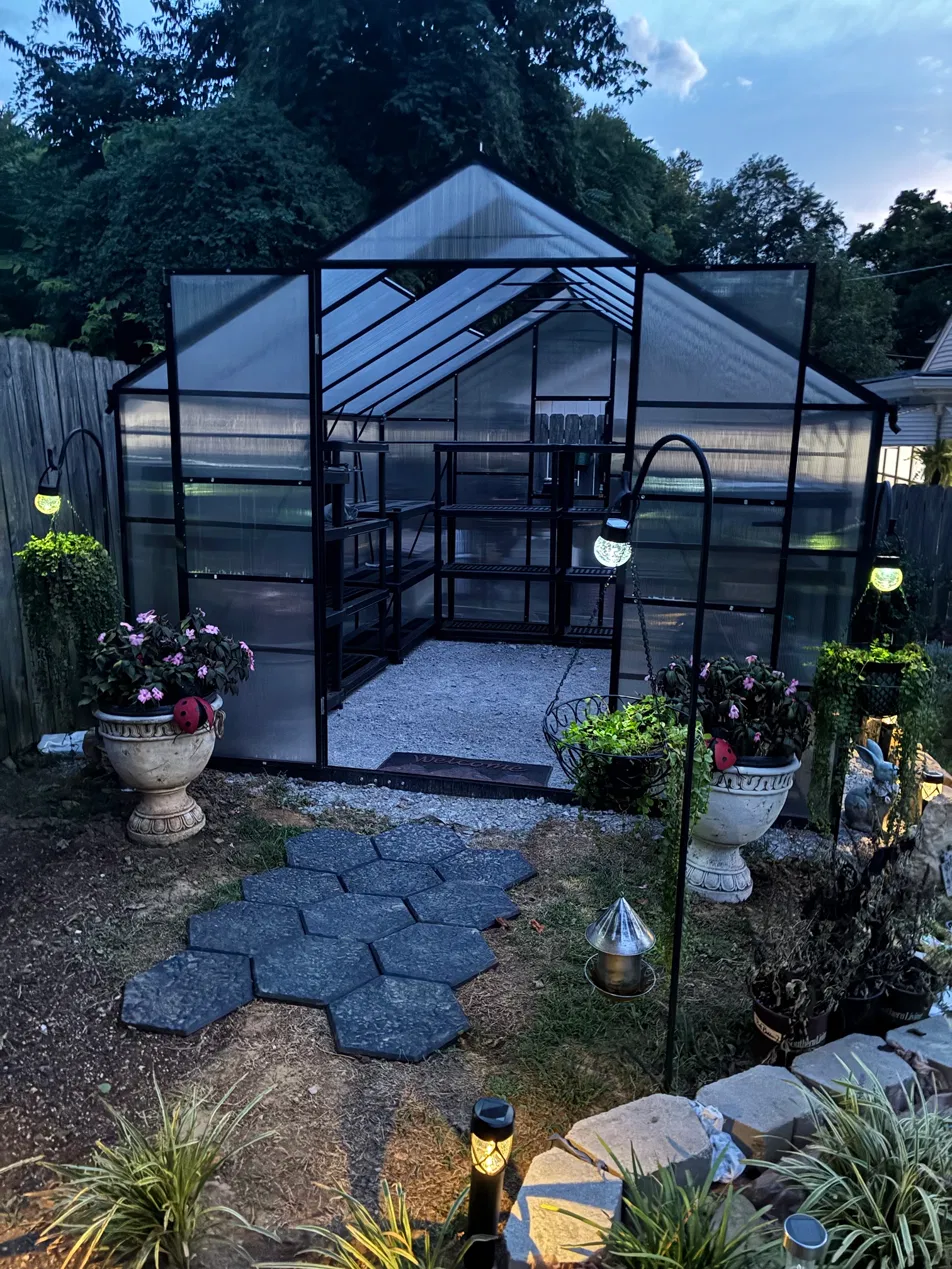 Marlaine’s black-framed greenhouse at dusk, lit from inside, with urns and lanterns out front